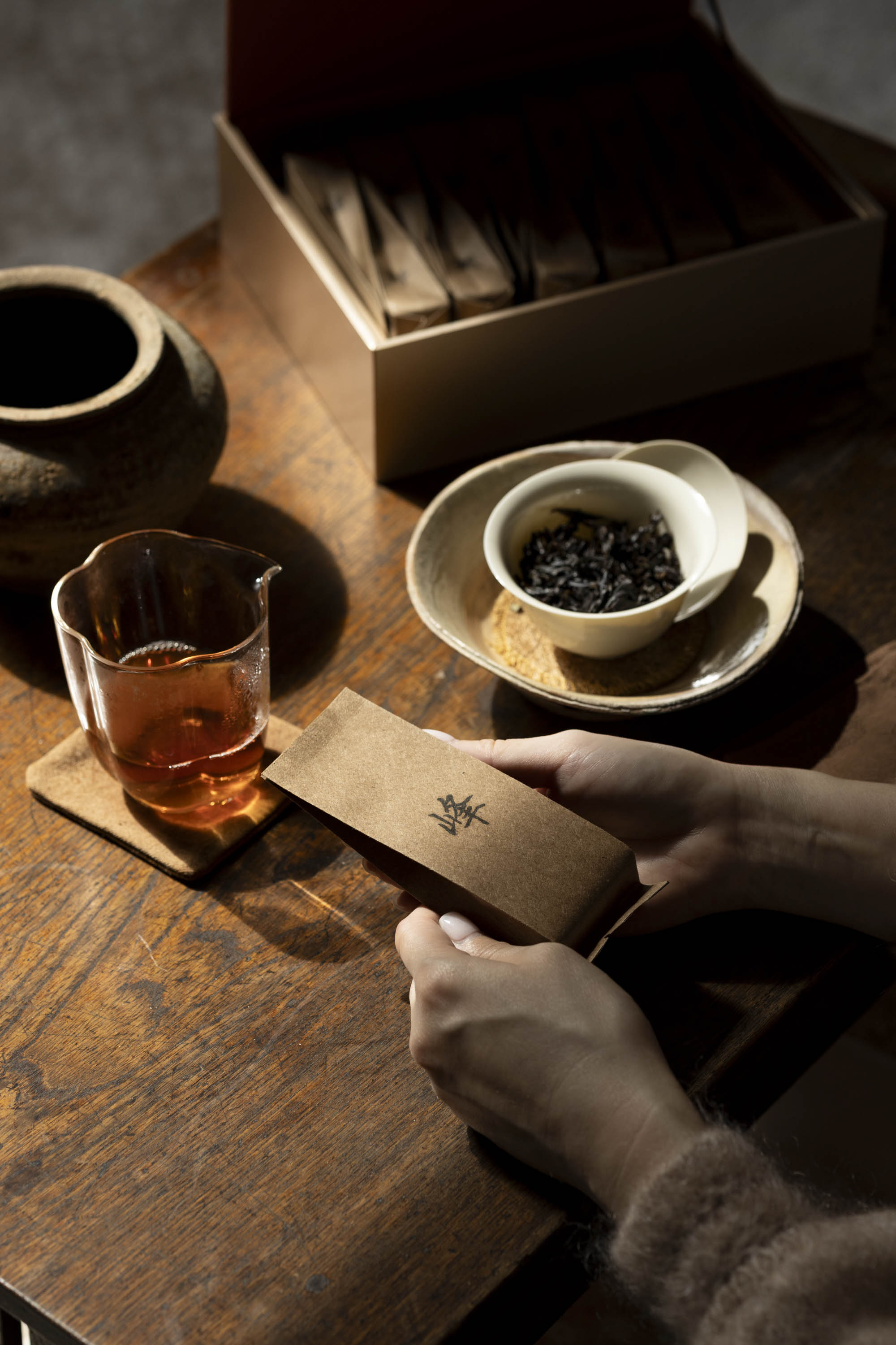A portion of Lian Hua Feng of Wuyi Yancha wulong tea next to a glass pitcher of brewed tea, a white gaiwan with dark leaves, and the open hyt box on a wooden table