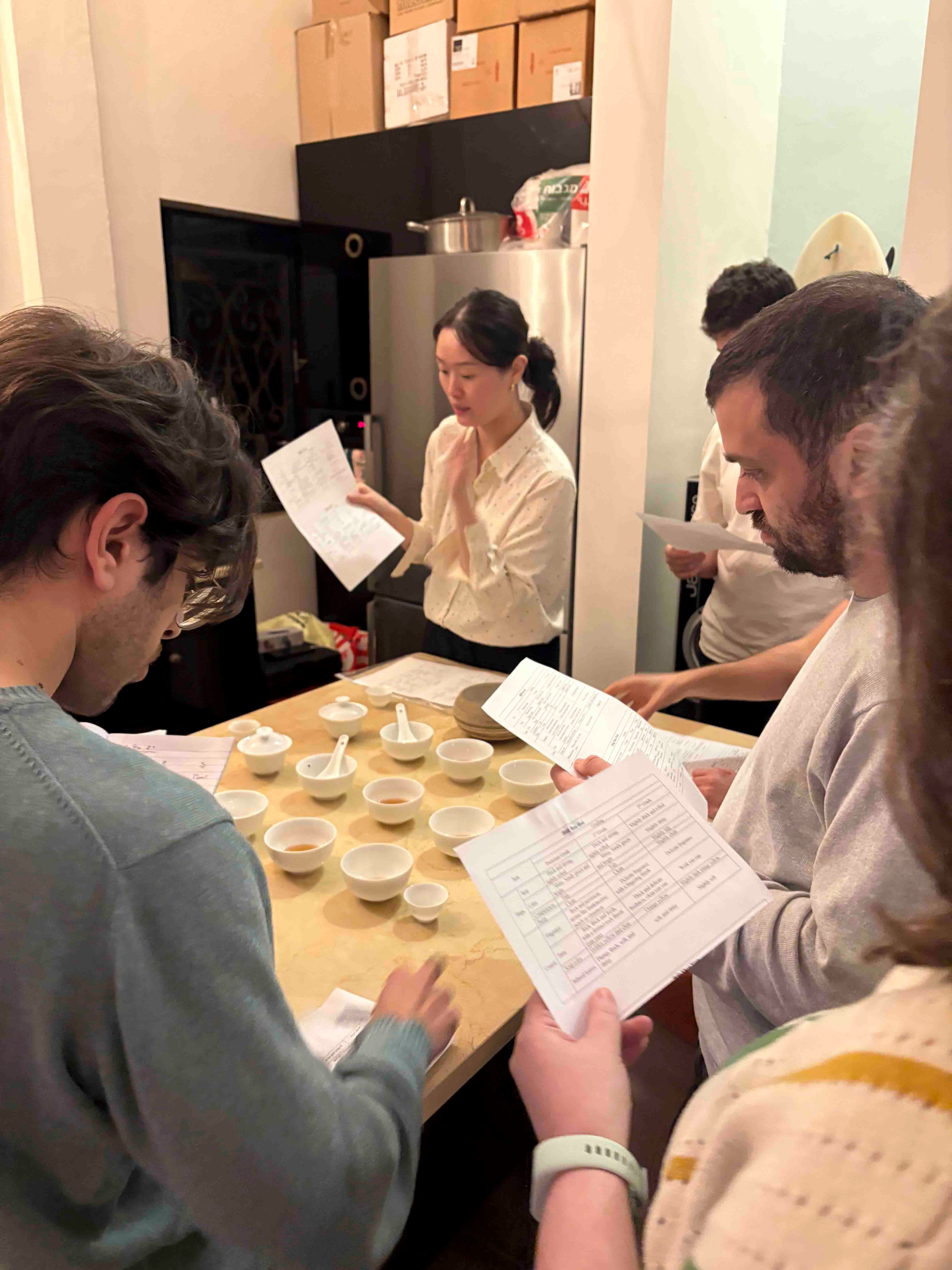 Pink Luk is teaching students the Chinese national standard evaluation for Wuyi Yancha wulong tea, with participants reading printed grading sheets around a table filled with white ceramic tasting bowls.