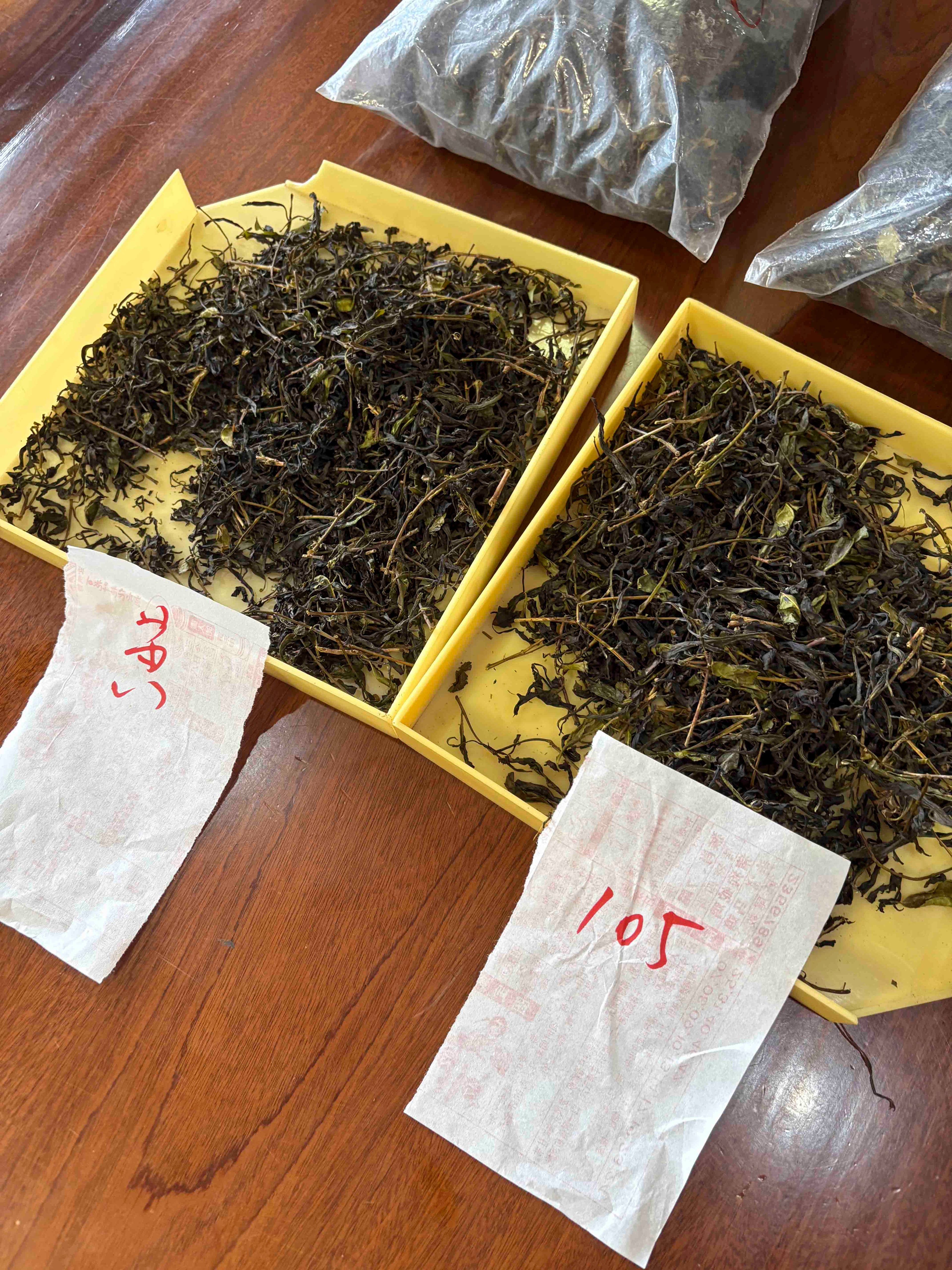 Two yellow trays filled with unfinished wulong tea leaves resting on a wooden table alongside handwritten paper labels during the sorting and evaluation process.
