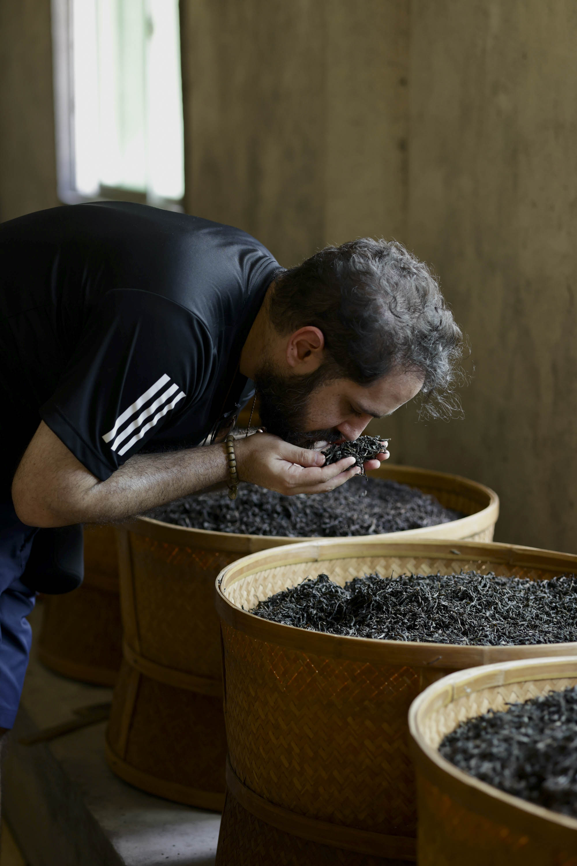 Nadi Biran smelling dark Wuyi wulong tea from his hands over woven bamboo baskets in a baking room at a tea factory.