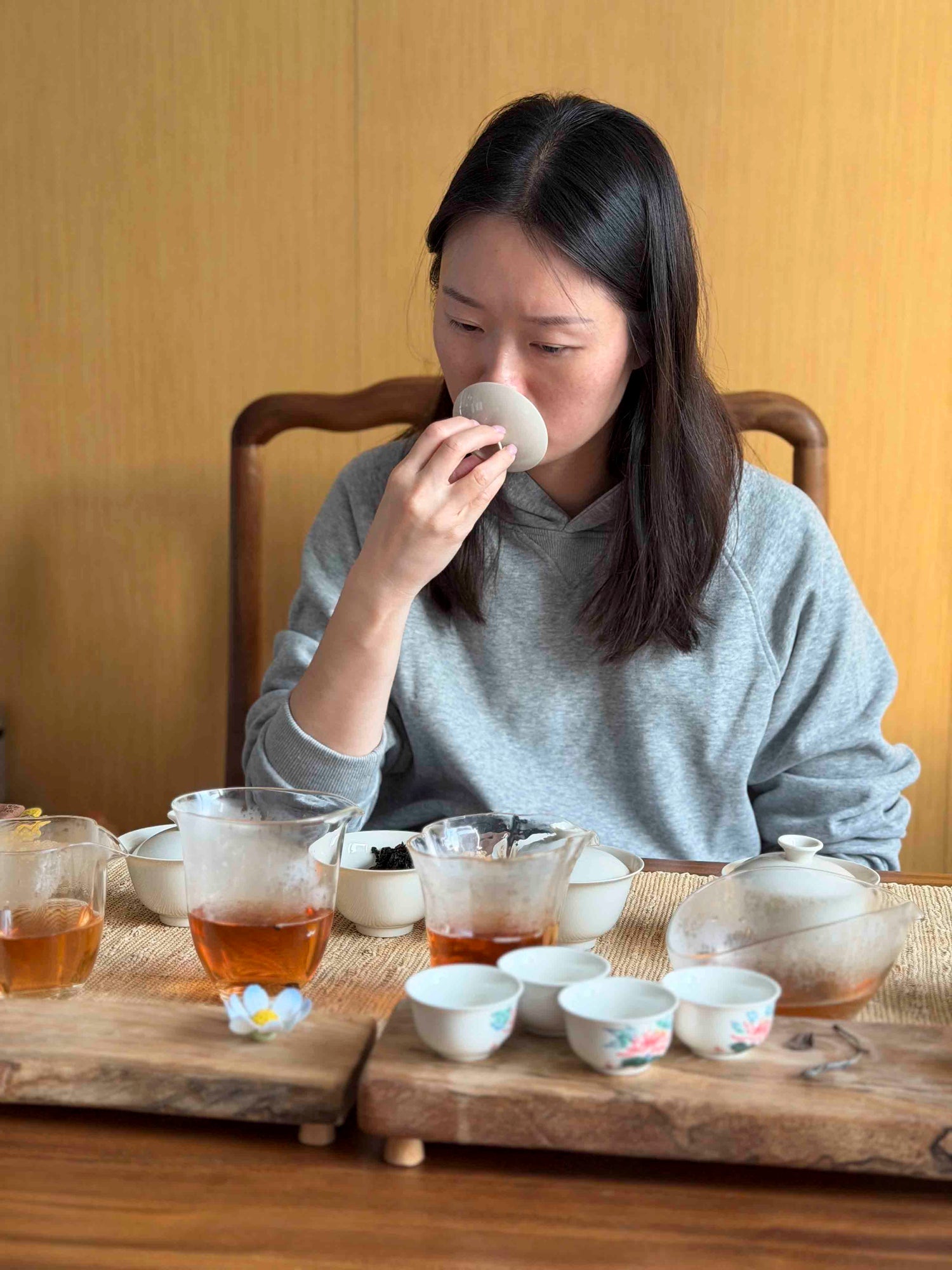 Pink Luk is evaluating the aroma of Wuyi Yancha wulong tea from a gaiwan lid during a tasting session, surrounded by glass pitchers and small ceramic cups on a wooden table.