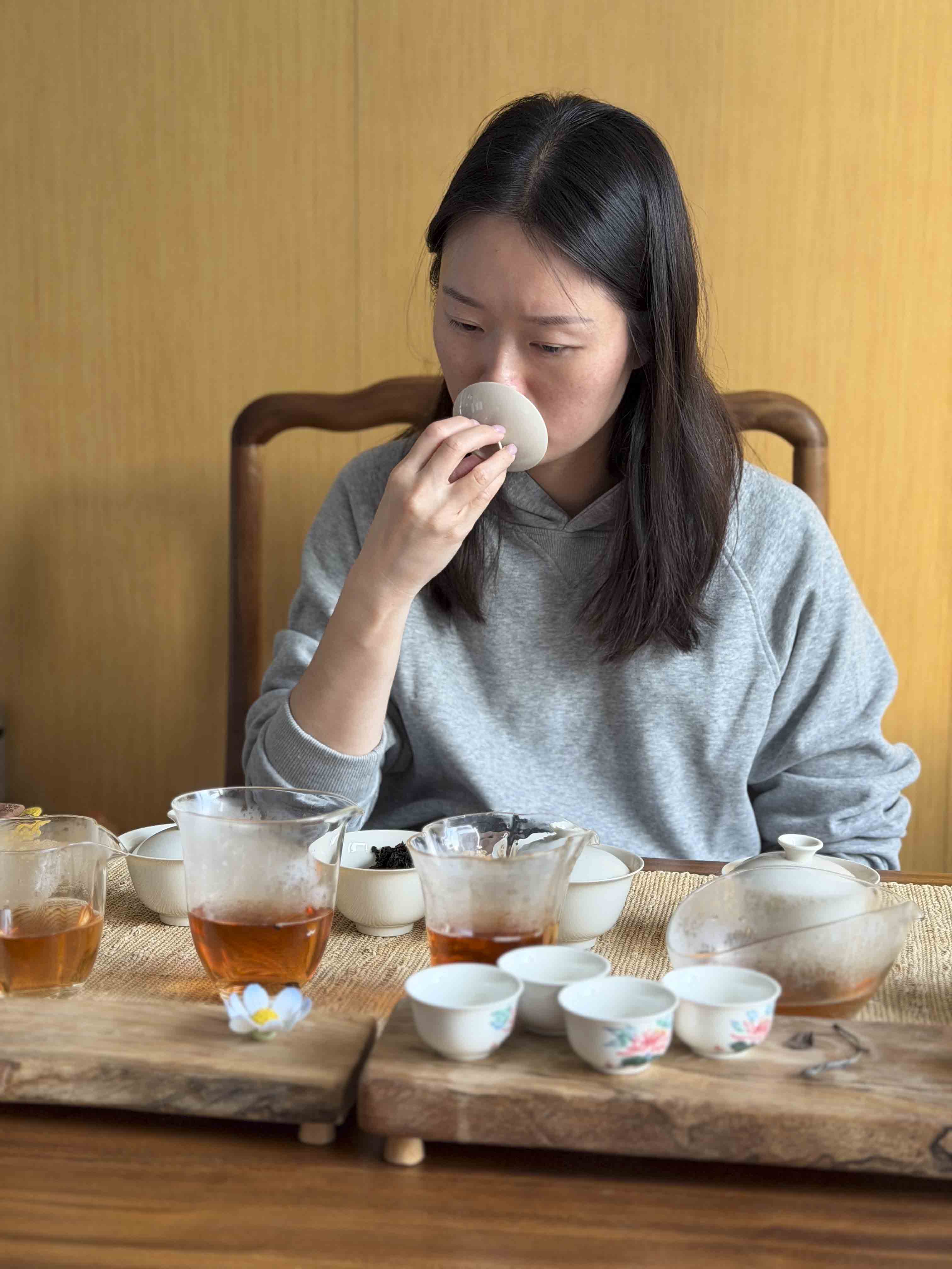 Pink Luk is evaluating the aroma of Wuyi Yancha wulong tea from a gaiwan lid during a tasting session, surrounded by glass pitchers and small ceramic cups on a wooden table.