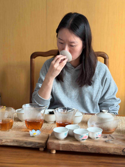 Pink Luk is evaluating the aroma of Wuyi Yancha wulong tea from a gaiwan lid during a tasting session, surrounded by glass pitchers and small ceramic cups on a wooden table.