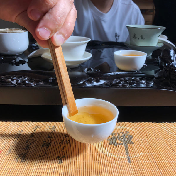 A hand holding a bamboo tea scoop above a white tasting cup of golden tea on a tea tray